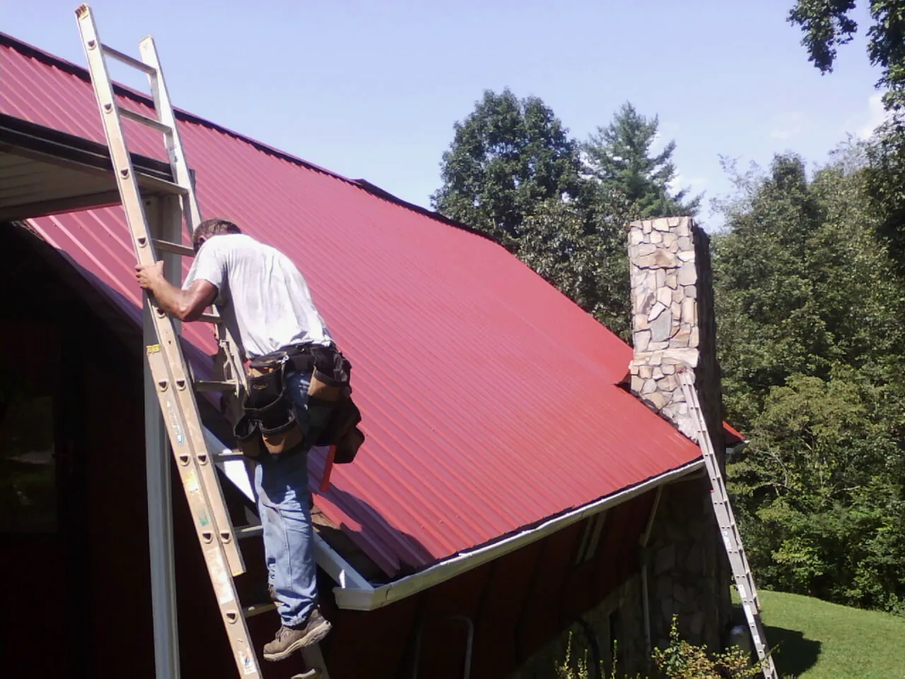 Roofer on a ladder beside a red metal roof and stone chimney — Soffit & Fascia Repair by Roofing Brothers in Phelan